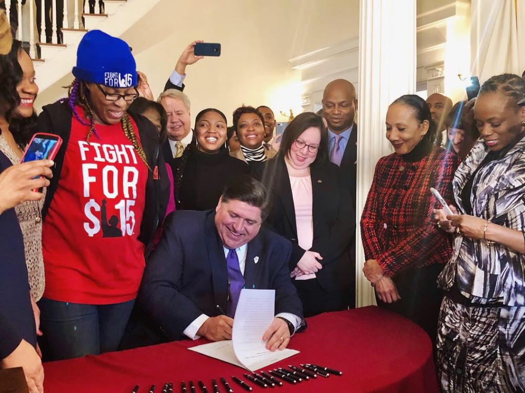 Feb. 19th, 2019, Gov. J.B. Pritzker signs historic bill to raise Illinois minimum wage to $15 an hour by 2025. Standing behind him is Tichina Haywood, a CNA earning $12.67 an hour whom Pritkzer said he would recognize her when he signed the bill. 